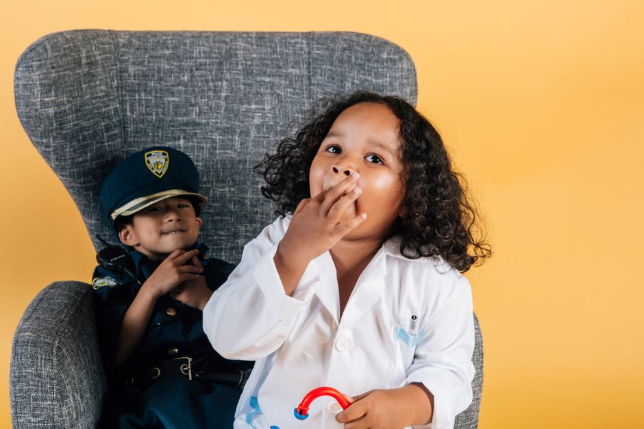 Asian boy in policeman costume and curly African American girl in doctor uniform sitting in armchair together while playing in studio on yellow background