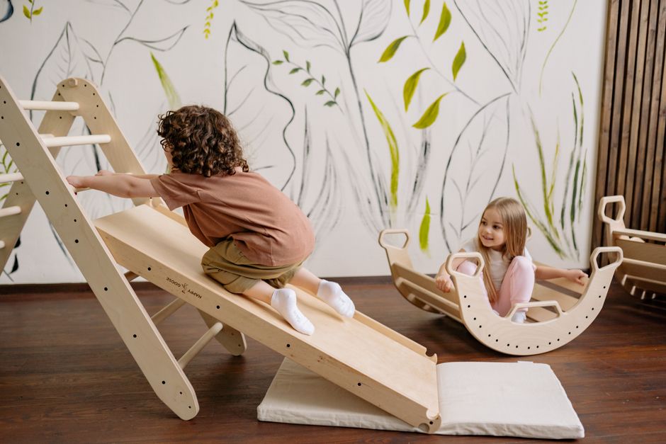 Two children playing indoors with a wooden slide and rocking toy in a playroom.