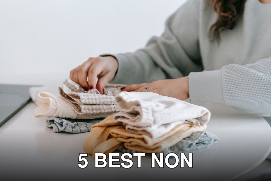 Crop faceless female in casual clothes folded colorful baby garments while sitting at white table in room on white background