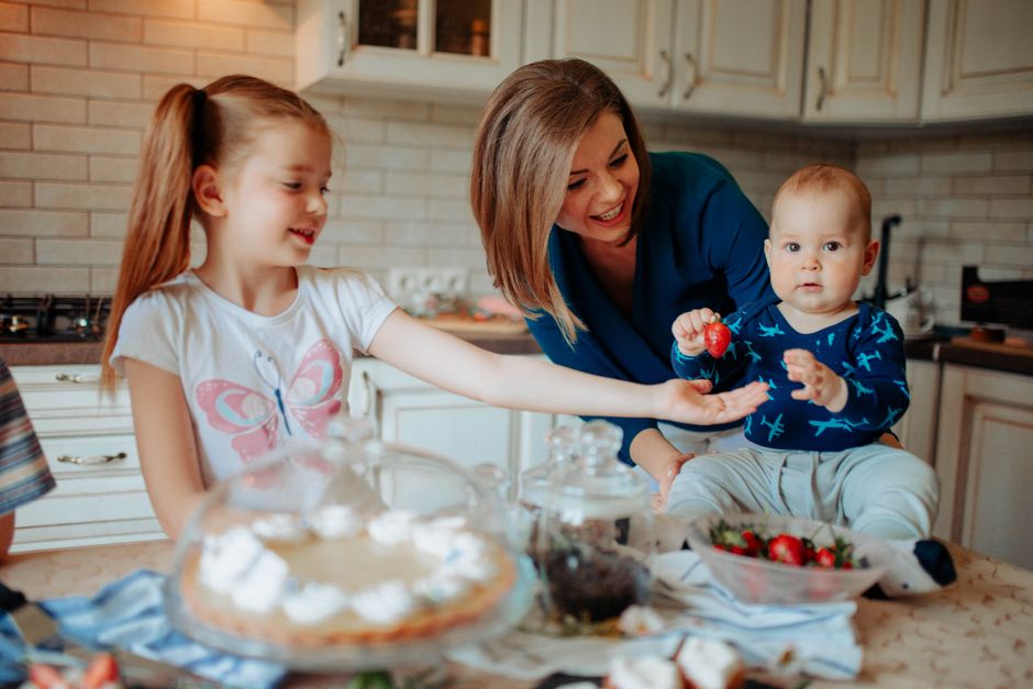 Children with mother in casual outfit standing near counter with strawberry and desserts with cream in bright kitchen near cabinets