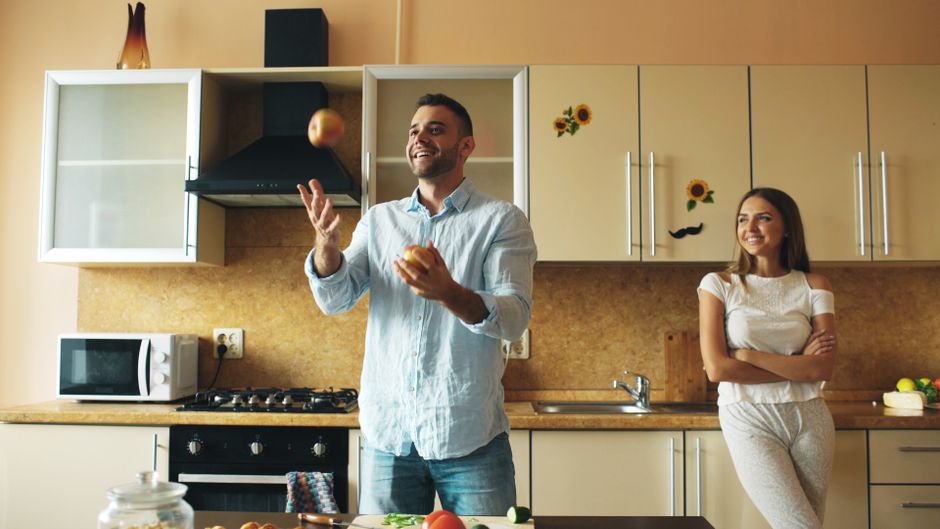 A cheerful man juggles apples in a bright, modern kitchen while a smiling woman watches.
