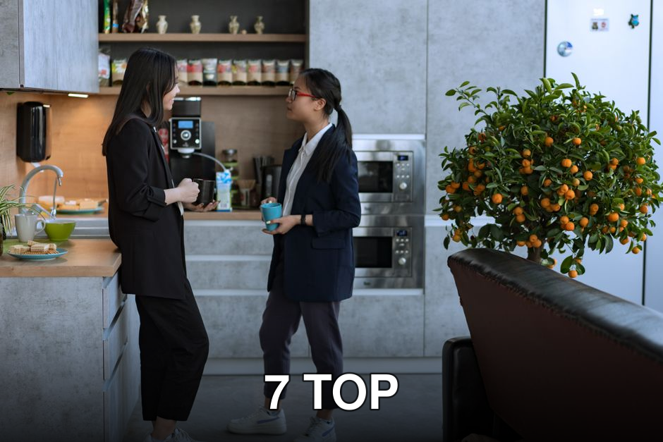 Two women chatting in a stylish office kitchen, holding mugs and enjoying a break.