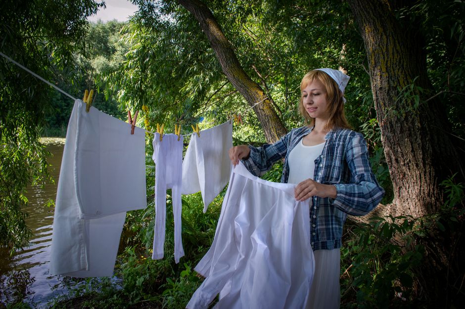 A woman hangs white laundry on a clothesline in a lush forest beside a river.