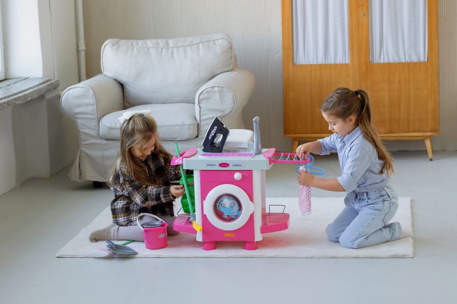 Two girls engage in imaginative play with a toy washing machine set in a cozy indoor setting.