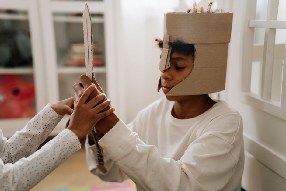A young child dressed in a cardboard helmet engages in imaginative play with a handmade sword indoors.