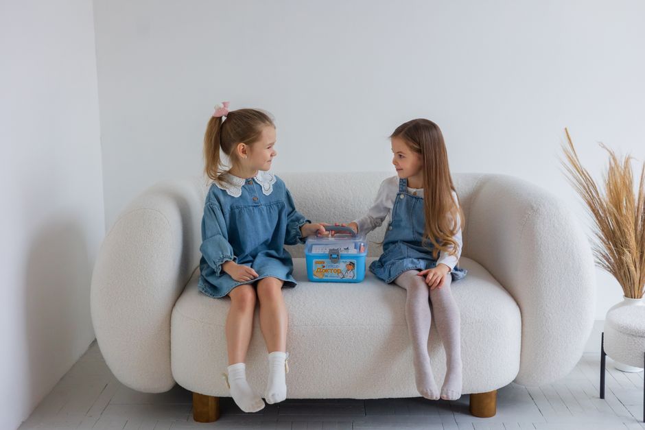 Two young girls sitting on a sofa, engaging with a toy doctor kit, showcasing imaginative play indoors.