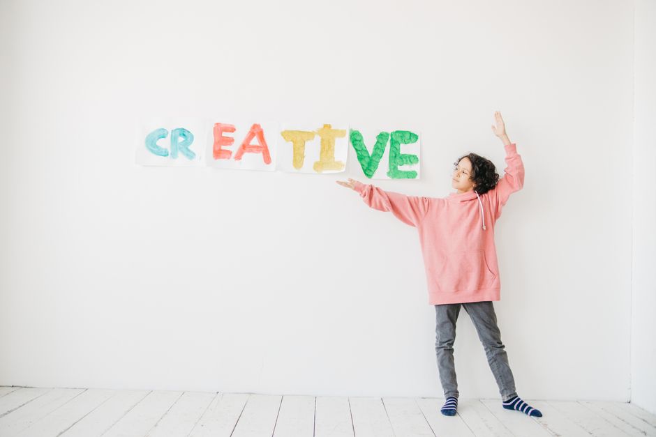 A child with curly hair in a pink hoodie points to colorful 'CREATIVE' text on a white wall.