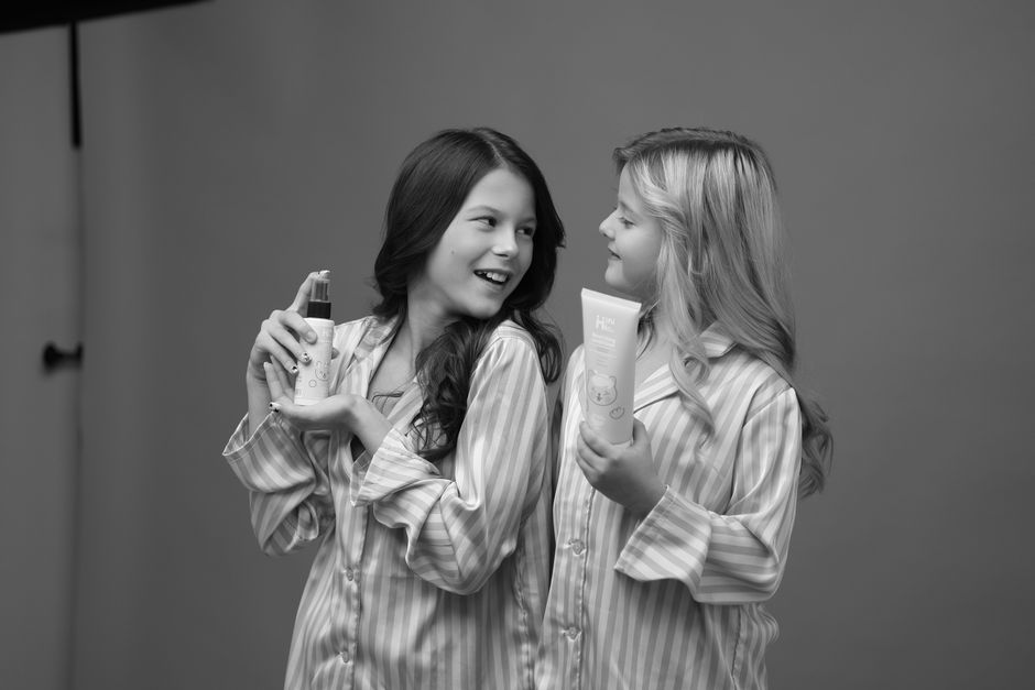 Two children smiling while holding skincare bottles, in matching pajamas.
