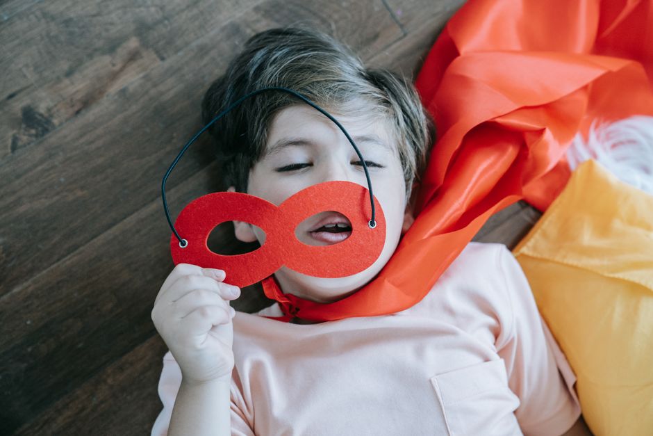 A child playfully holding a red eye mask while lying down indoors, expressing imagination and joy.