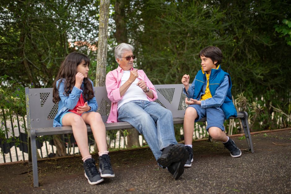 A grandfather sits with his grandchildren on a park bench, surrounded by nature, enjoying a sunny day.