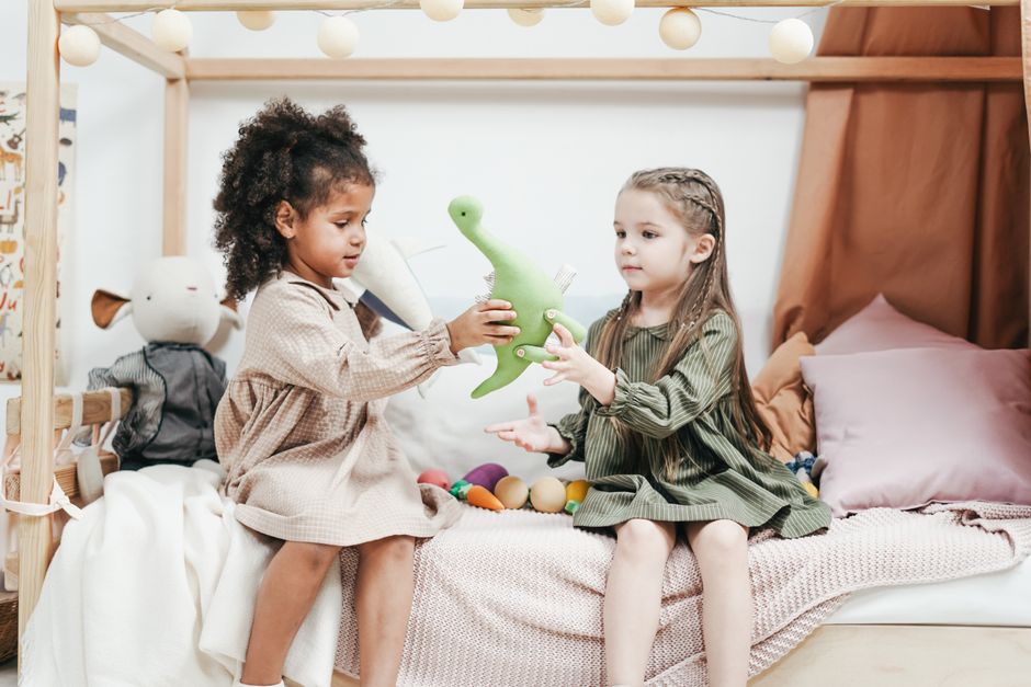 Two young girls sharing a plush dinosaur while sitting on a bed in a cozy playroom.