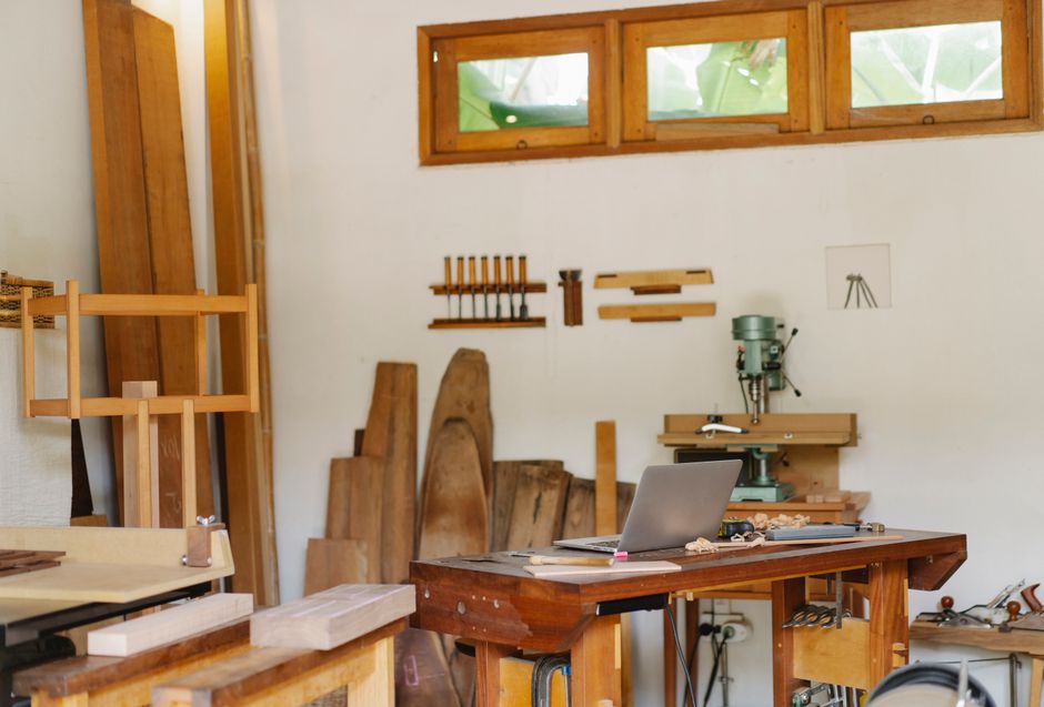 Opened laptop placed on table in traditional carpentry workshop full of wooden boards and tools in sunlight