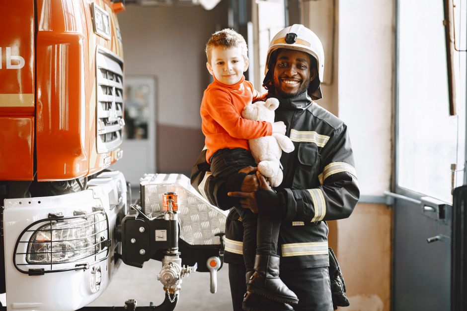 A smiling firefighter holds a boy with a teddy bear at the fire station.
