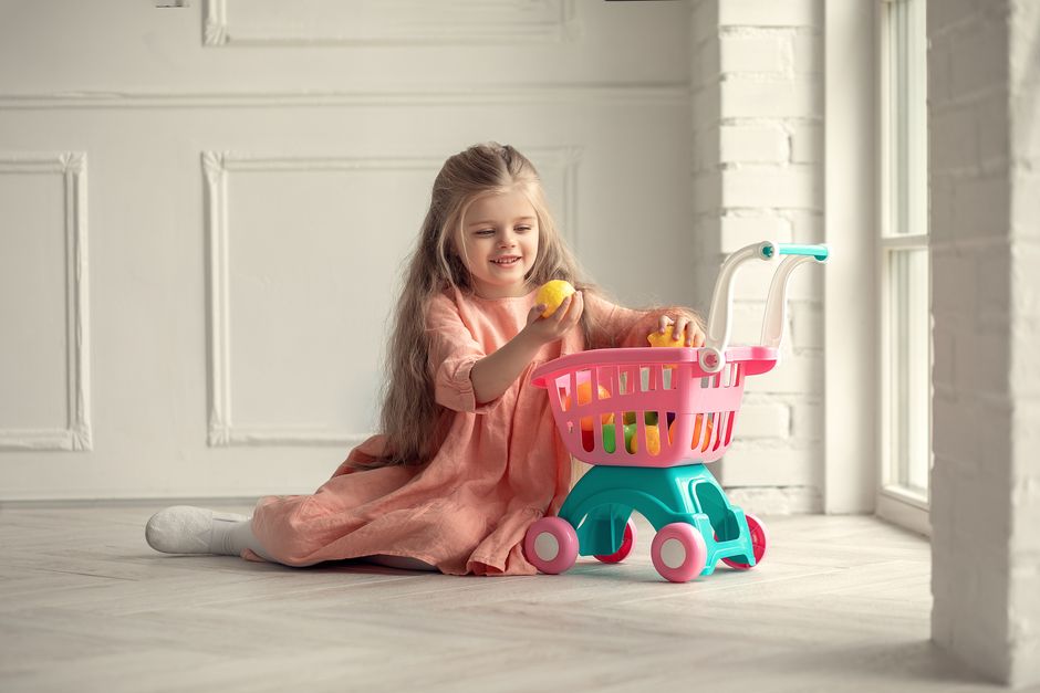 A joyful child with long hair plays with a colorful toy shopping cart indoors.
