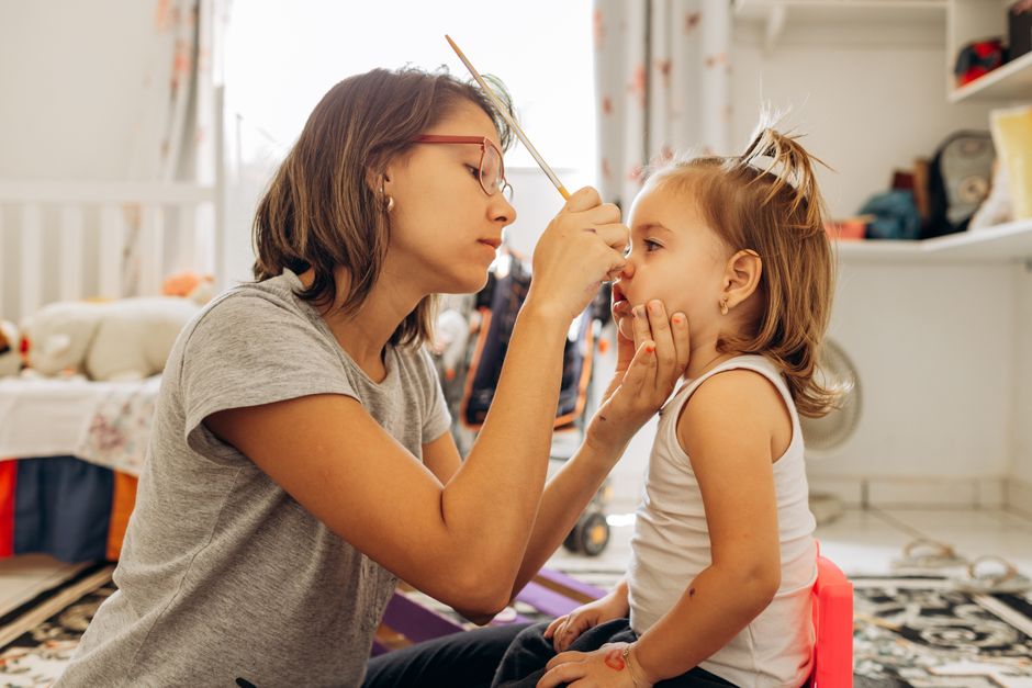 A mother meticulously face-paints her young daughter in a cozy playroom.