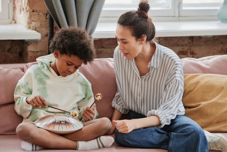 A boy playing a steel tongue drum with guidance from a female instructor on a sofa.