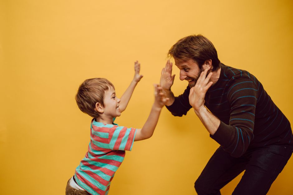 Father and son dressed casually, joyfully sharing a high five against a vibrant yellow background.