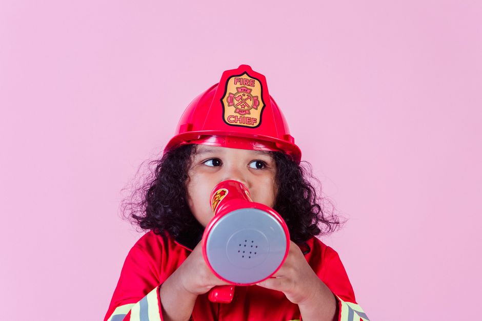 Cute little African American girl wearing bright firefighter costume and helmet and talking in toy loudspeaker while playing in light studio and looking away