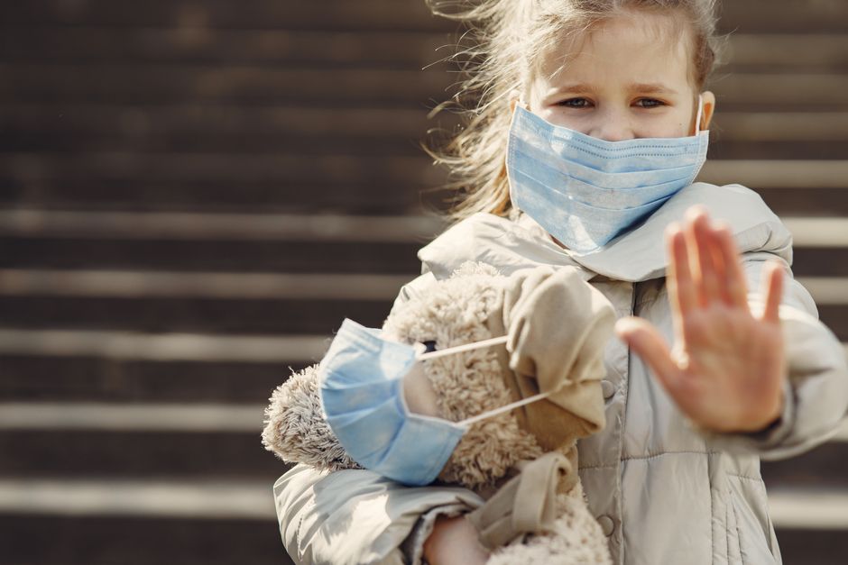 Worried child wearing warm clothes and mask hugging teddy bear in medical mask and showing stop gesture while looking at camera