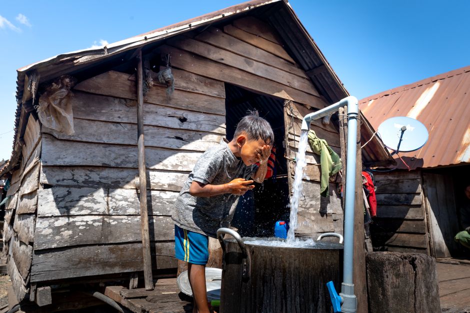 A young boy washes his face at an outdoor tap beside a wooden hut in a rural area.