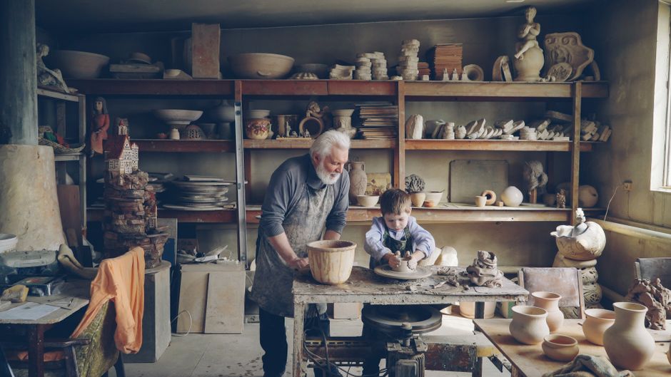 A grandfather teaches his grandson pottery in a cozy studio filled with ceramics.