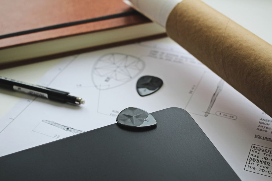 Close-up of guitar pick design on technical drawings and black notebook.