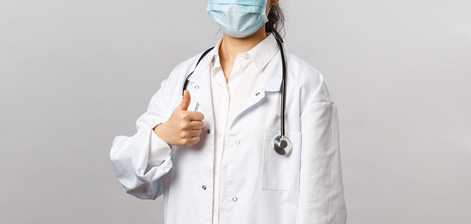 A confident doctor wearing a face mask and giving a thumbs up in a medical setting.
