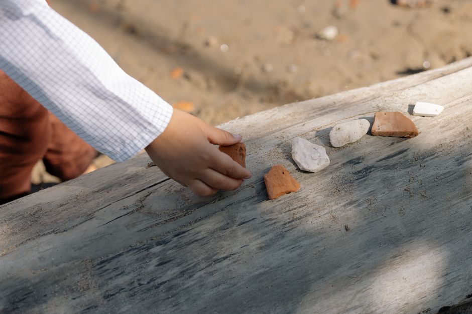 A child's hand arranges various stones on a piece of driftwood in an outdoor setting.
