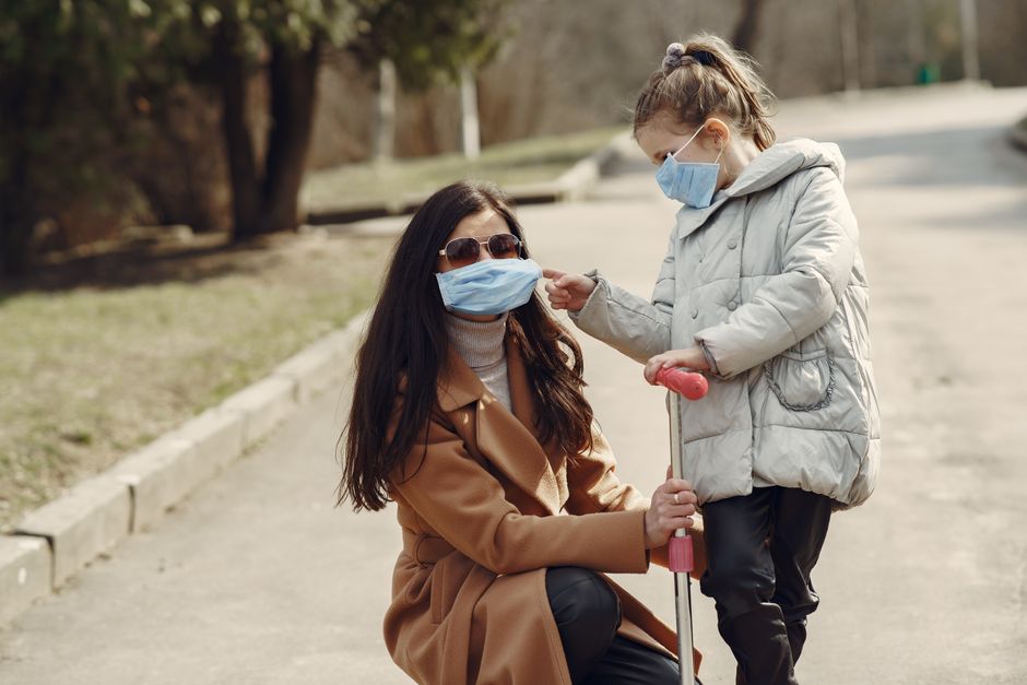 Young female in warm clothes and sunglasses squatting and supporting scooter of daughter while girl helping mom to put on mask