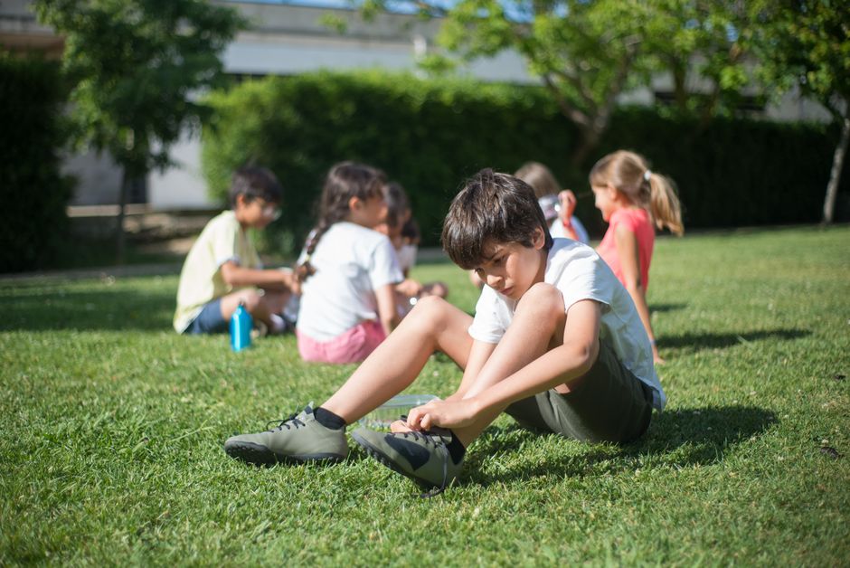 A child tying shoelaces while sitting on grass in a sunny park surrounded by friends.