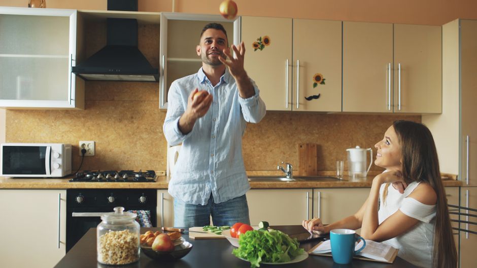 A man juggling apples while a woman watches in a modern kitchen setting.