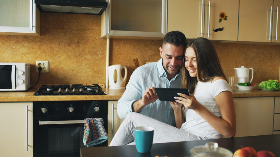 Happy couple using a tablet in a modern kitchen setting, surrounded by cozy and warm decor.