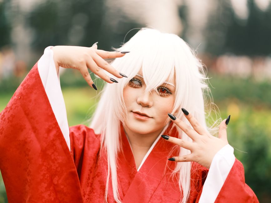 Person in cosplay with red robe and white wig posed outdoors in Vietnam garden setting.
