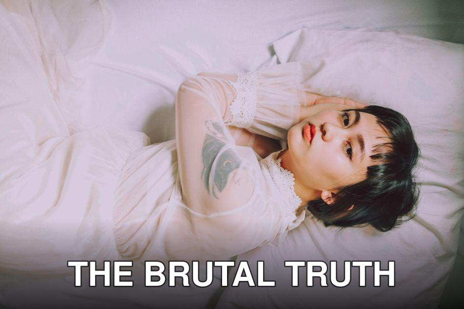 Stylish woman with tattoo posing on a bed in soft lighting, capturing a sense of elegance and beauty.