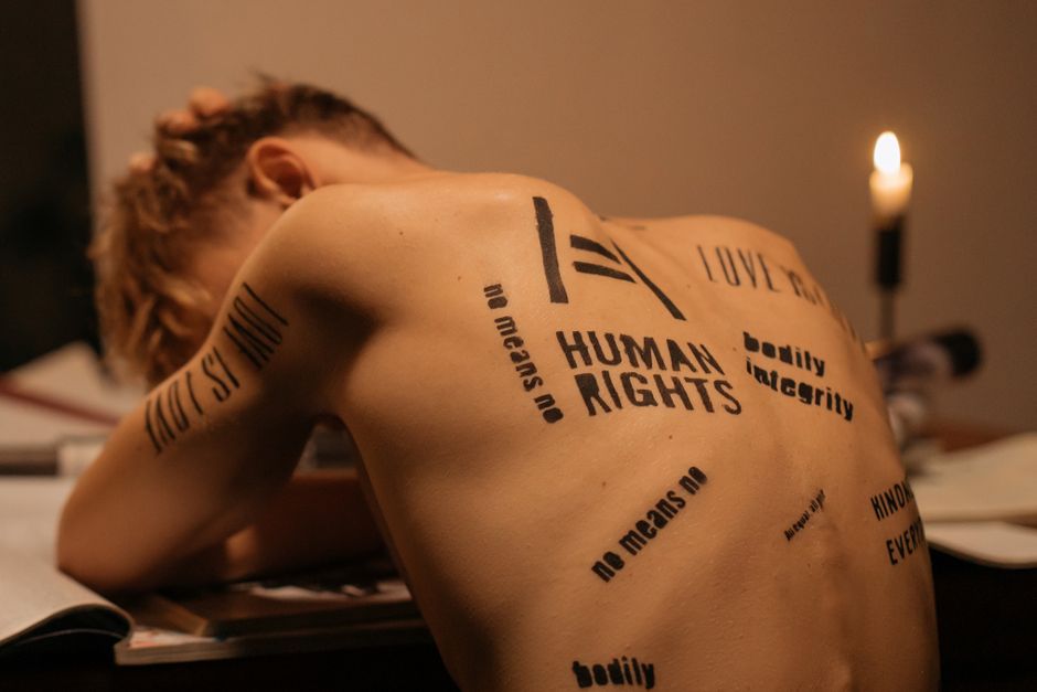 A shirtless man with messages on his back supporting human rights in a dimly lit room.