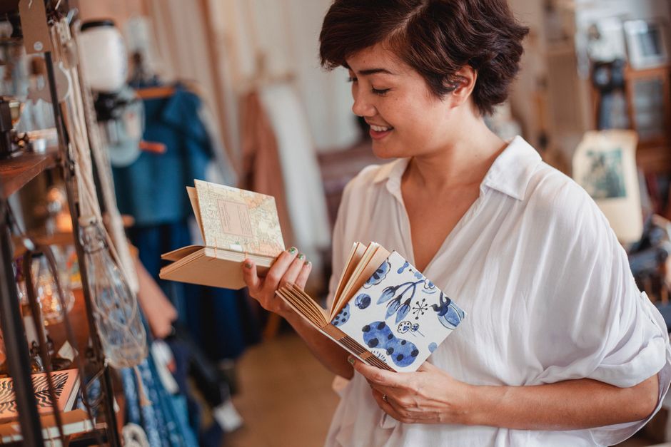 Side view of crop young happy Asian lady choosing various notepads while standing near shelf with stylish goods in vintage boutique