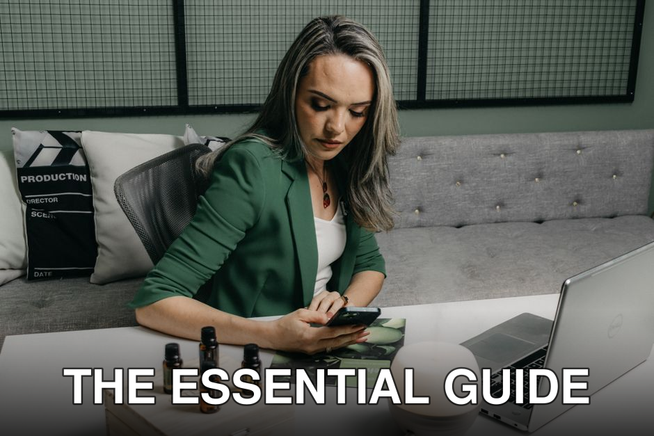 Focused businesswoman using smartphone at desk with essential oils and laptop, blending work and wellness.