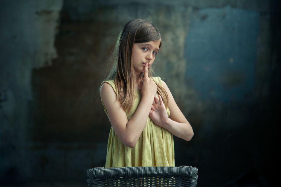 A young girl in a yellow dress gestures for silence with a finger on her lips, standing against a moody background.