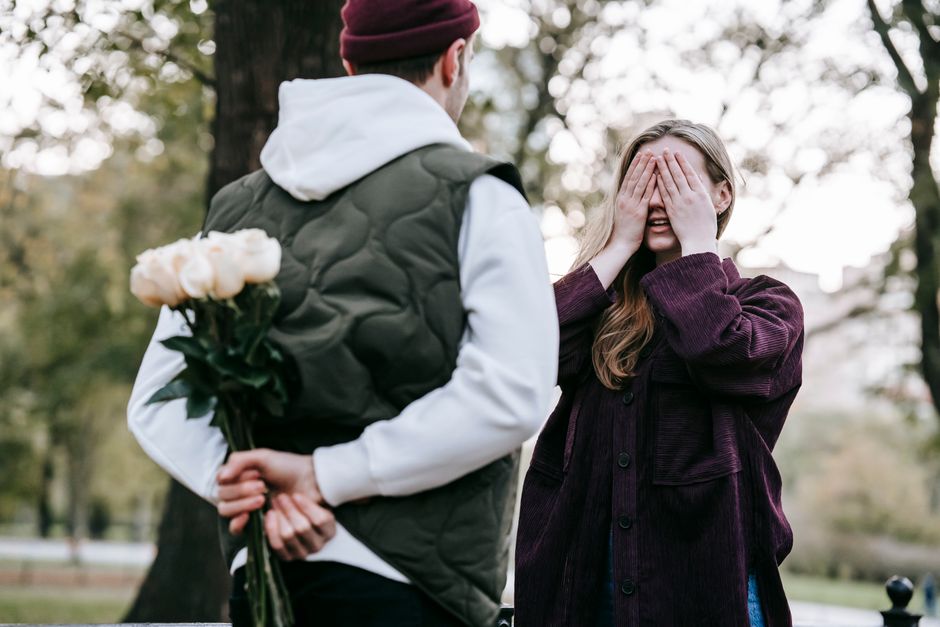 Loving young male in hat and warm clothes going to give bunch of flowers to girlfriend covering eyes with hands while having date in park
