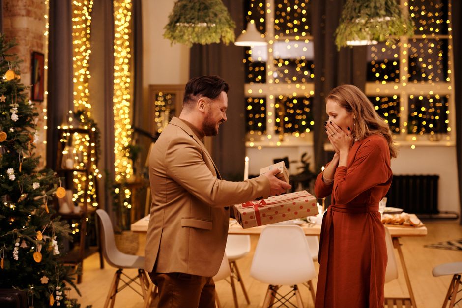 A couple exchanging Christmas gifts in a warmly decorated living room with festive lights.
