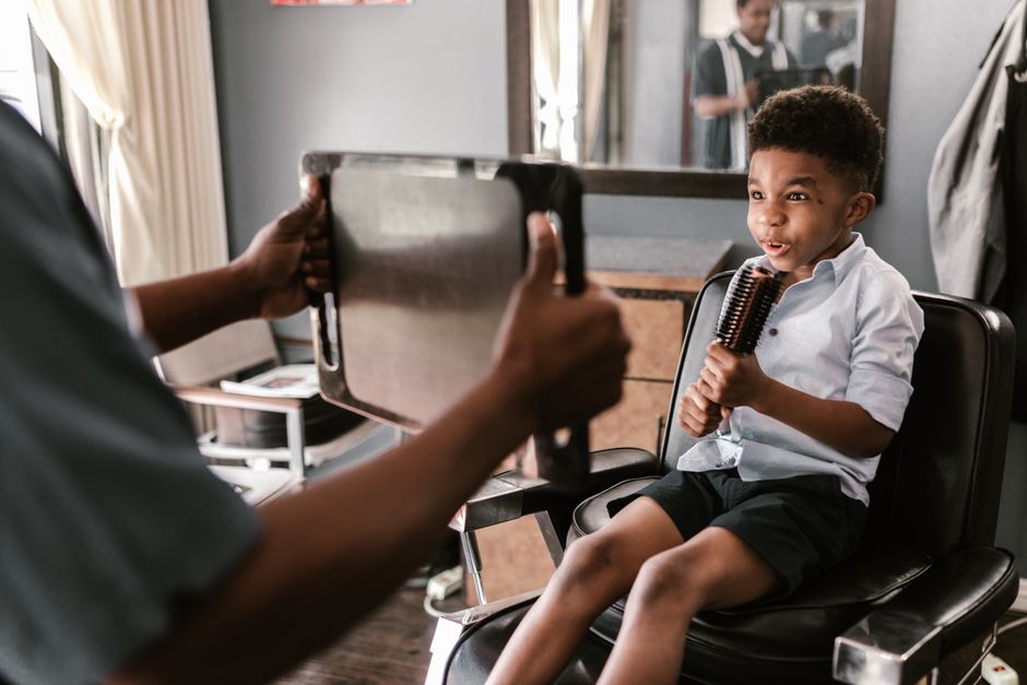 A joyful young boy sits in a barber chair holding a brush during a haircut indoors.