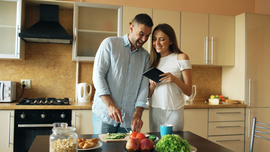 Smiling couple preparing a meal, chopping vegetables and using a tablet in a cozy kitchen setting.