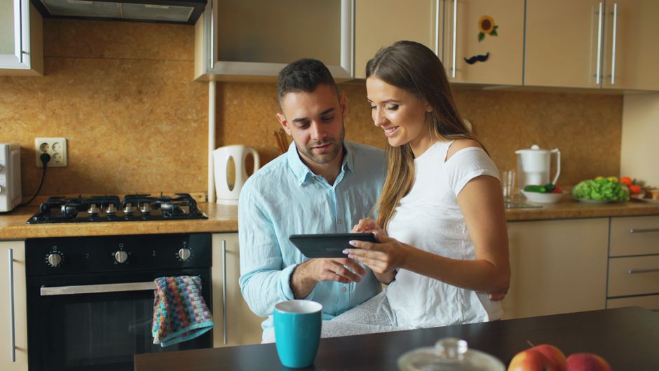 Happy couple using a tablet in a cozy kitchen setting, discussing a recipe.