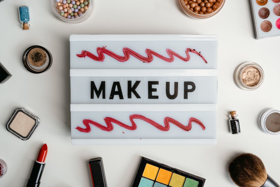 Flat lay of makeup items with lipstick, powder, and palette surrounding a sign reading 'MAKEUP'.
