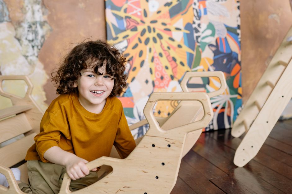 Smiling child with curly hair enjoying playtime on wooden toy indoors with colorful background.