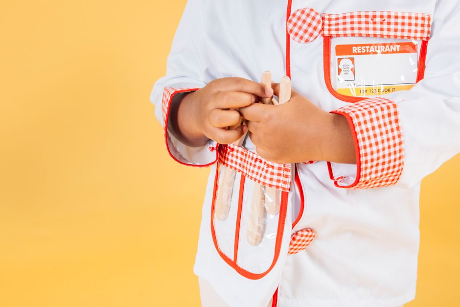 Crop anonymous ethnic kid with supplies for kitchen wearing chef jacket in studio against yellow background