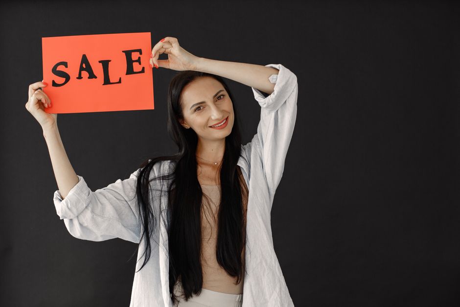 A cheerful woman with long hair in casual attire holds a red sign with 'SALE' written on it.